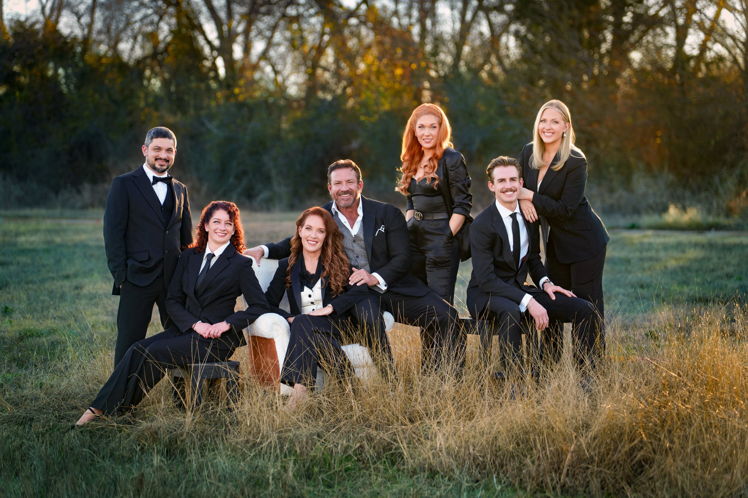 Extended family in formal black attire gathered in a field of tall grass at sunset