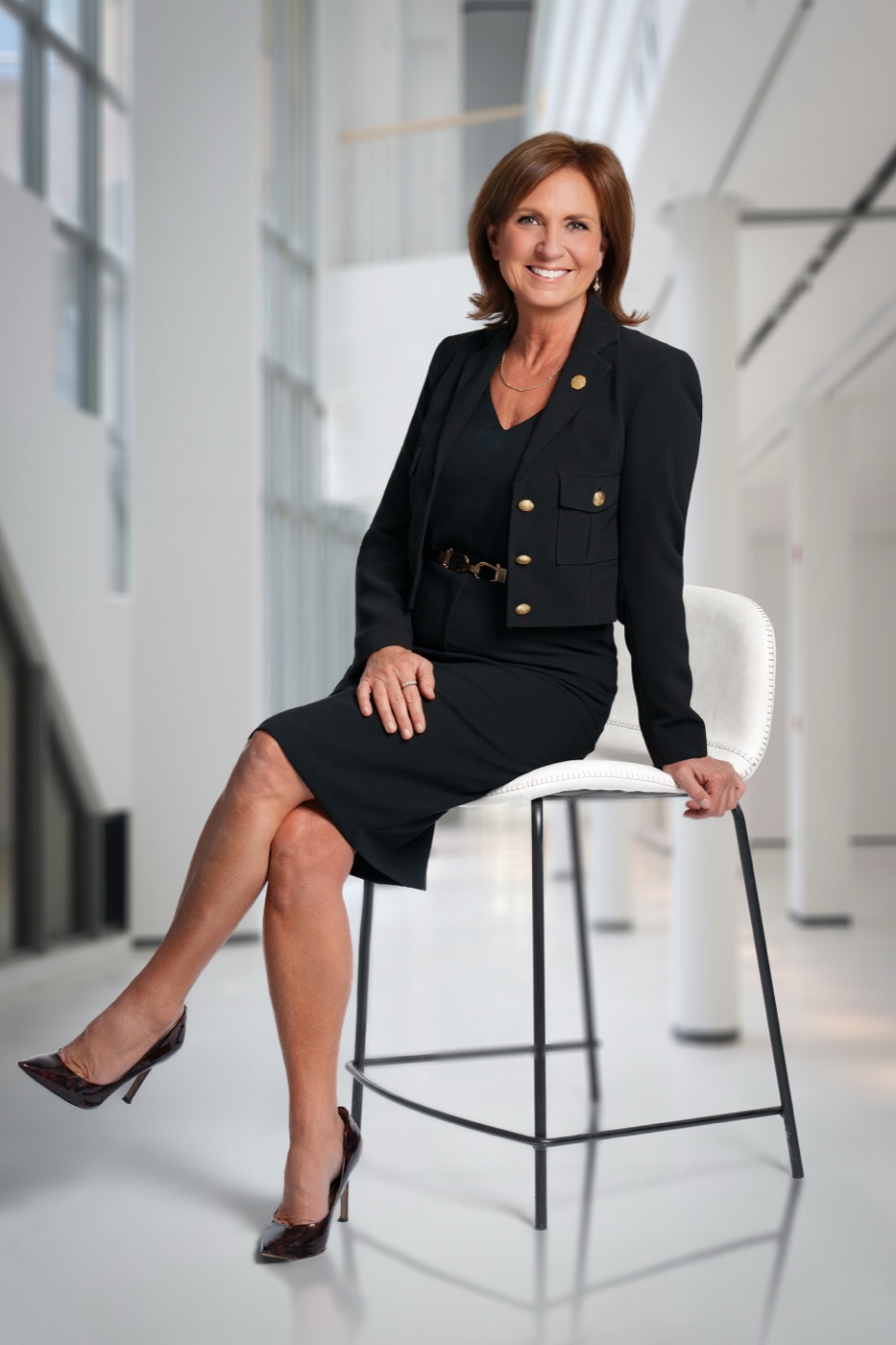 Studio portrait of a businesswoman in a black suit seated on a stool with a bright office atrium background added
