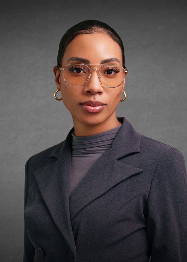 Studio headshot of a woman in glasses and a charcoal blazer on a grey background